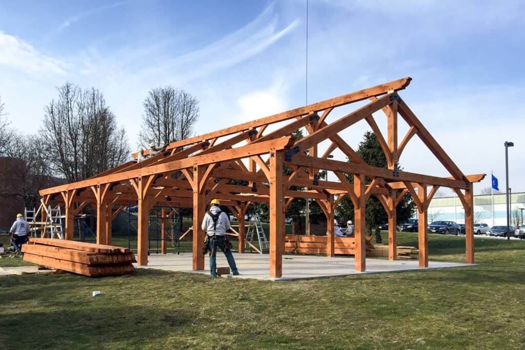 Several workers are assembling the wooden frame of a large open-sided pavilion on a concrete slab in a grassy area, with piles of wood nearby and trees and buildings in the background.