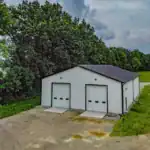 Aerial view of white steel workshop with black trim and twin overhead garage doors, built by Swiss Construction in Sunbury, Ohio, set against a wooded backdrop and open field.