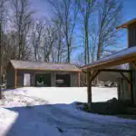 Metal garage with open bays housing compact construction equipment, surrounded by snow and woods near Walhonding, Ohio.