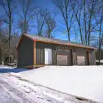 Three-bay post-frame garage with dark board-and-batten siding and brown garage doors, set in a rural snowy clearing.