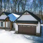 Snow-covered wooded lot surrounding the West Howard Residence, a modern farmhouse with dark roof, timber accents, and board-and-batten siding.