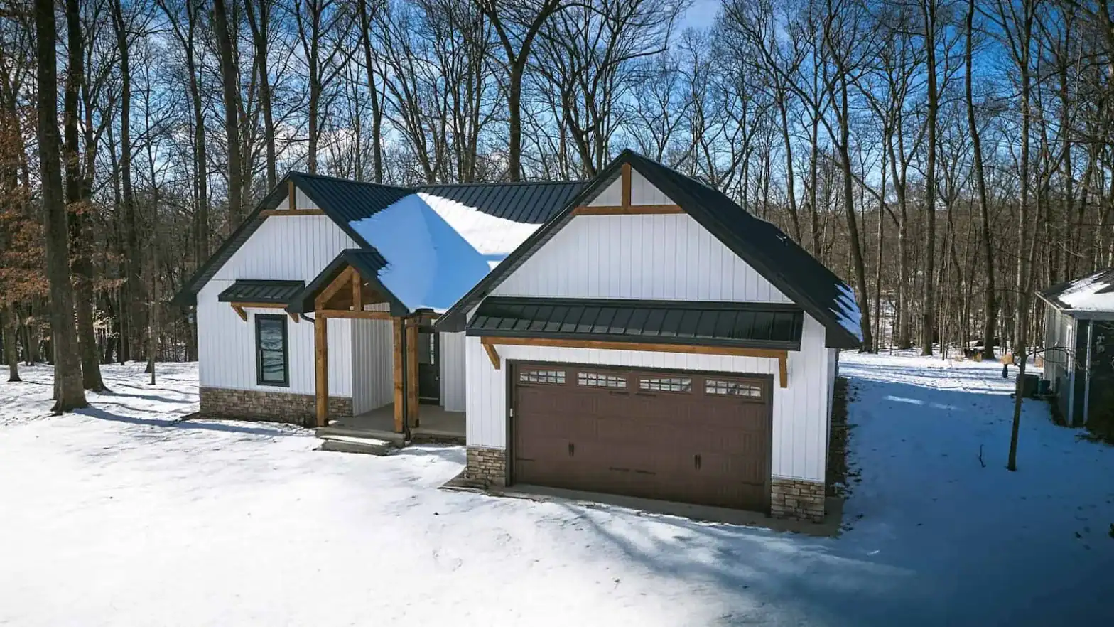Snow-covered wooded lot surrounding the West Howard Residence, a modern farmhouse with dark roof, timber accents, and board-and-batten siding.
