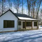 Rear view of the West Howard Residence featuring black-framed windows, white siding, and timber porch posts set against a winter forest backdrop.
