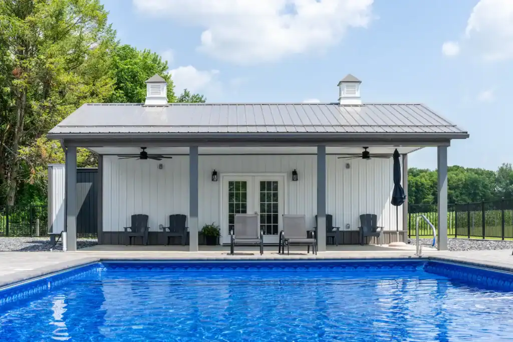 White pool house with gray metal roof, cupolas, and patio furniture overlooking a blue swimming pool