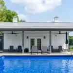 White pool house with gray metal roof, cupolas, and patio furniture overlooking a blue swimming pool