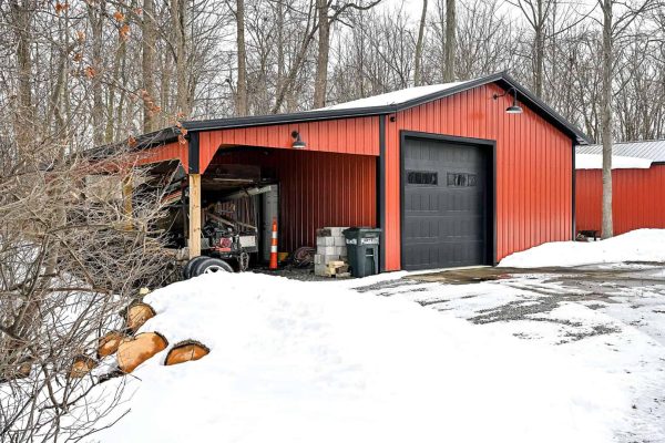 Auxiliary red metal outbuilding with garage bay and open lean-to storage area surrounded by snow, located at Columbia Station hobby property near Cleveland, OH.